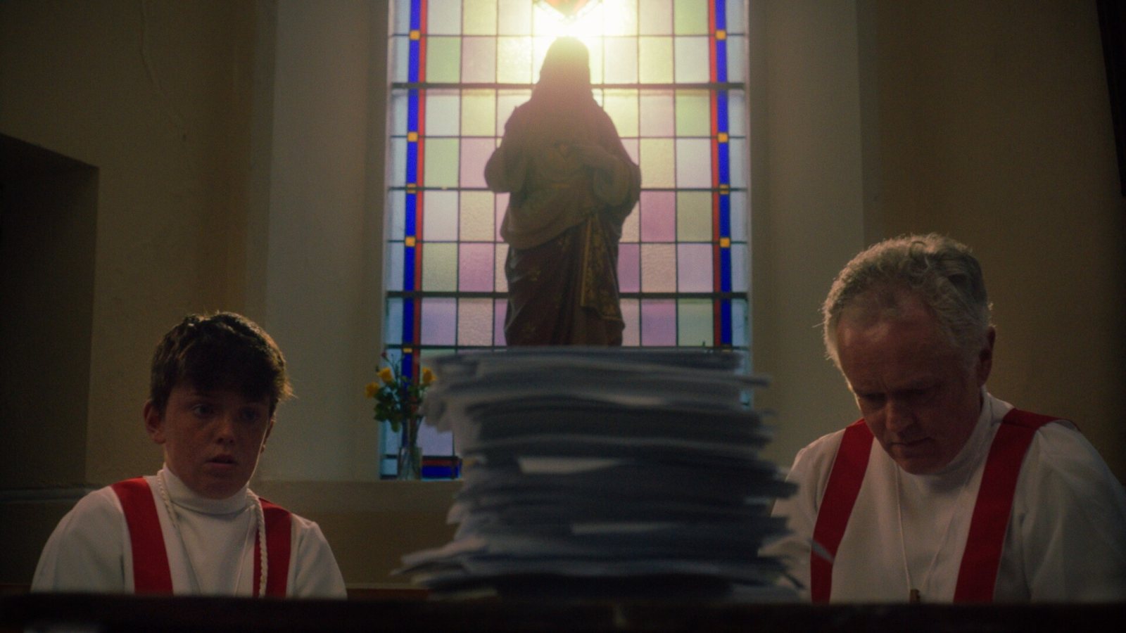 Still from Ireland's Oldest Altar Boy: a boy and an older man in altar robes beneath a stained-glass window