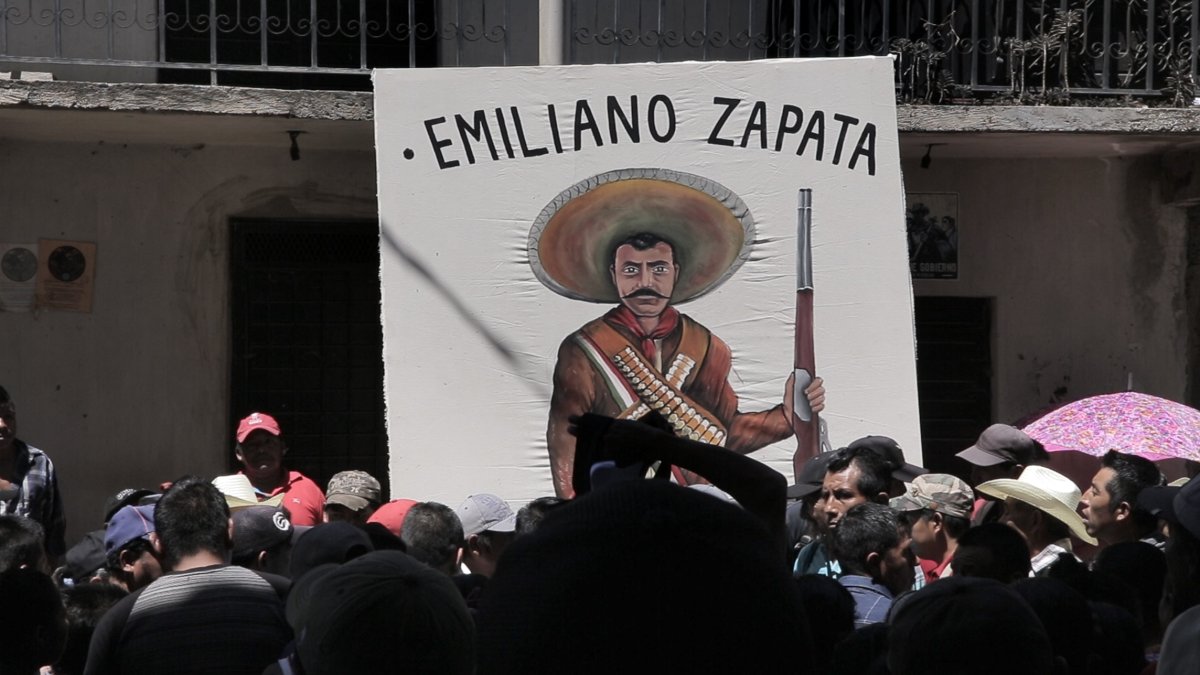 Still from A Bigger Place: a crowd gathered beneath a hand-painted banner of Emiliano Zapata